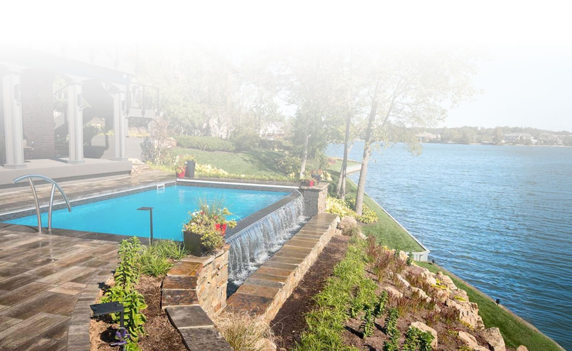 An in-ground backyard pool with a stone waterfall spillover overlooking a large lake on a sunny day.