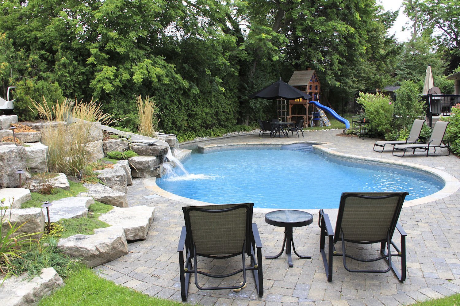 Backyard landscape featuring a freeform vinyl liner pool with a custom natural rock waterfall, pool slide, paver patio, patio loungers, and surrounding lush greenery.