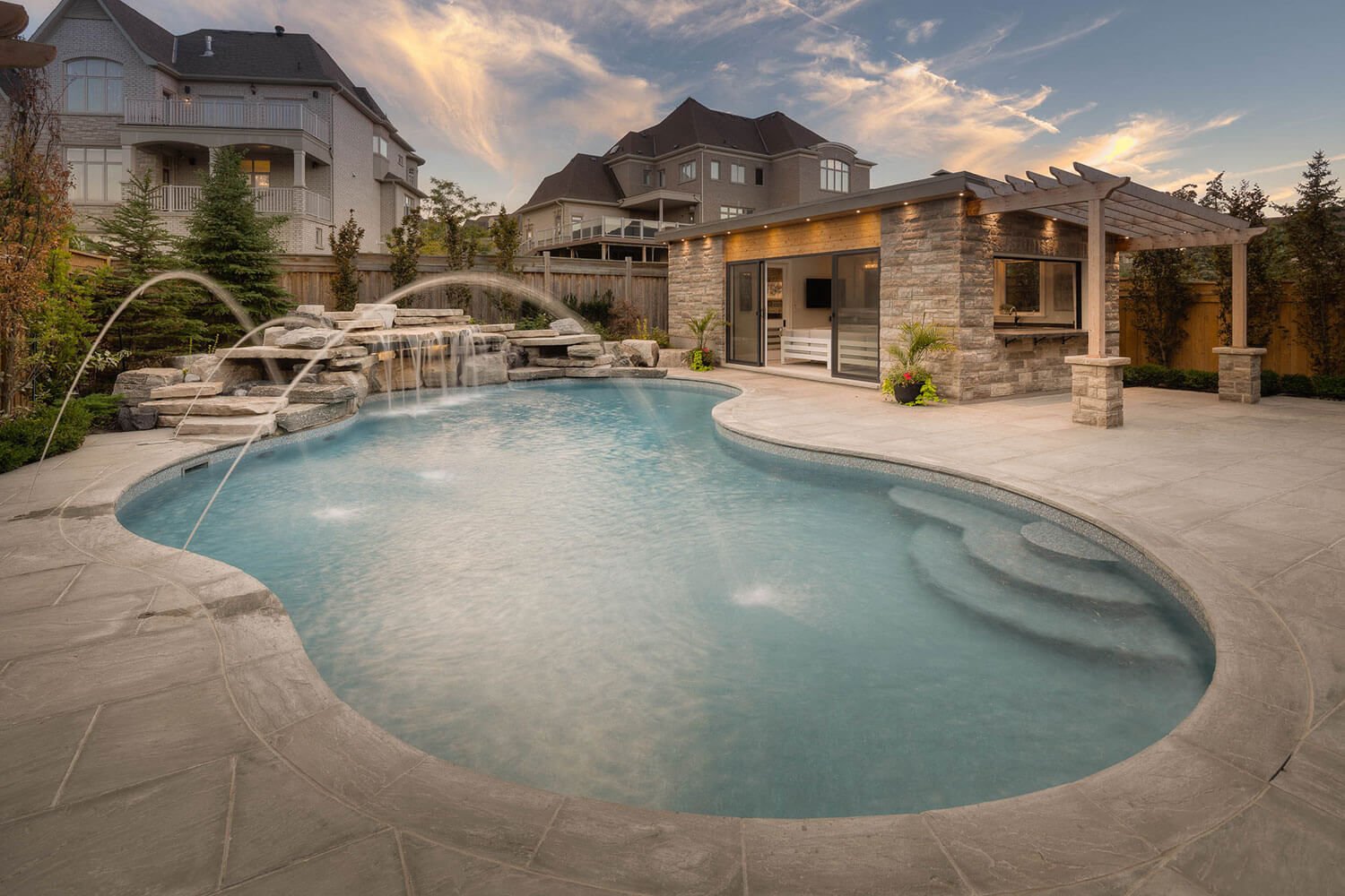 Daytime view of a freeform vinyl liner pool featuring a natural tiered rock waterfall, deck jet water streams, integrated entry steps, stone patio decking, and a modern cabana house.