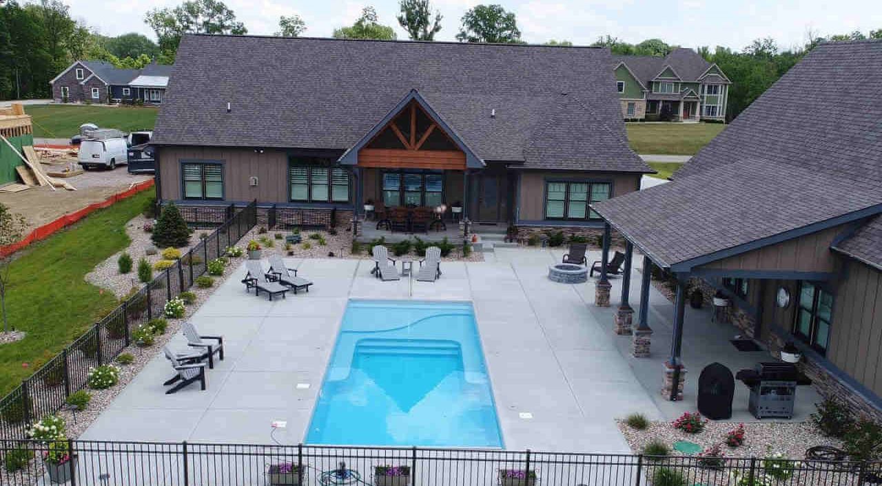 Elevated high-angle view of a rectangular fiberglass pool featuring an integrated tanning shelf and entry steps, surrounded by a spacious light concrete patio with lounge chairs, a fire pit area, black safety fencing, and a Craftsman-style home.