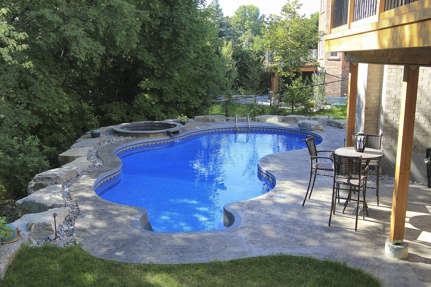 Backyard view of a freeform vinyl liner pool with bright blue patterned lining, stamped concrete decking, an elevated circular spillover hot tub, and surrounding natural rock landscaping.
