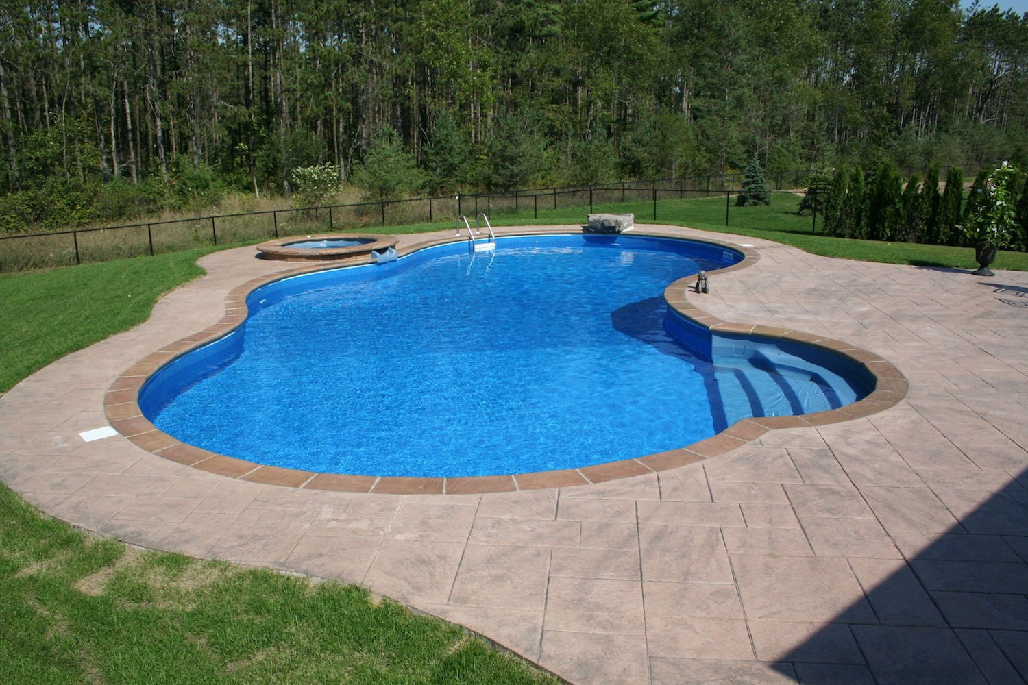 Wide daytime shot of a freeform vinyl liner pool with bright blue water, built-in curved entry steps, an integrated circular spillover hot tub, brown bullnose coping, and stamped concrete patio decking.