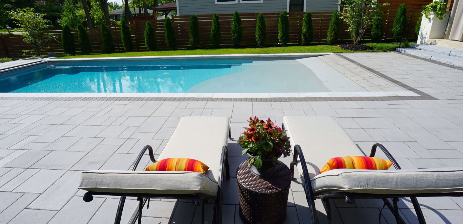 Side view of a rectangular fiberglass pool featuring a zero-entry beach slope design, surrounded by light gray paver decking with two chaise lounge chairs, a side table with flowers, and a wooden privacy fence with arborvitae trees in the background.