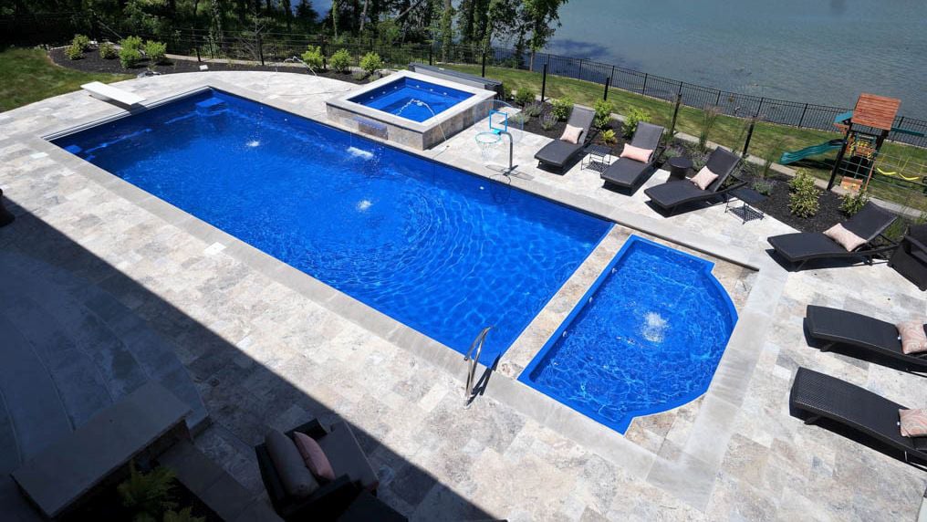 Aerial view of a luxury backyard installation featuring a large rectangular deep-blue fiberglass pool, an attached semicircular tanning ledge with water bubbler, an elevated square spillover spa, and surrounding stone paver patio overlooking a lake.