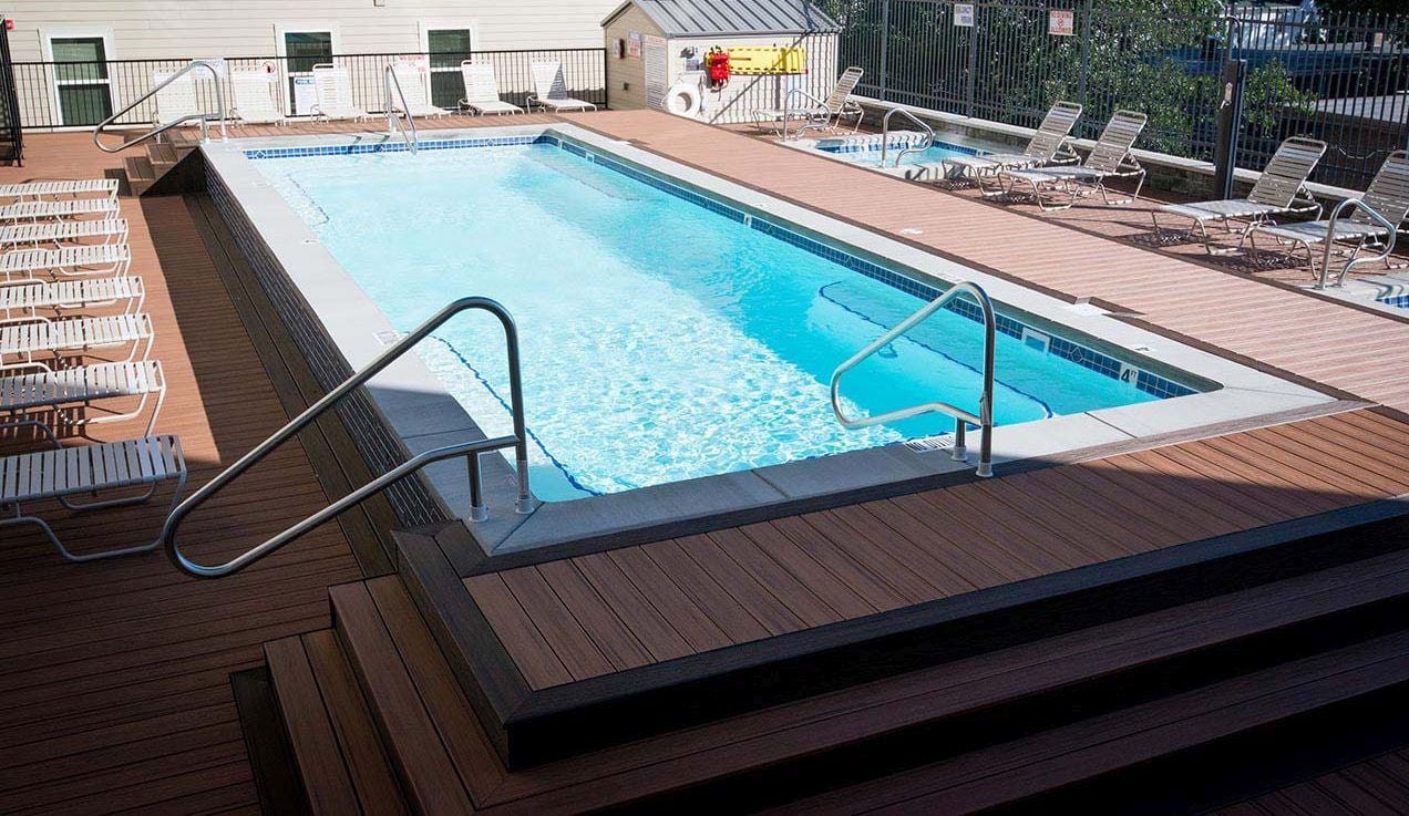 High-angle length perspective of a rectangular light-blue fiberglass pool featuring entry steps at both ends, dual stainless steel handrails, waterline tile, and an elevated composite wood patio deck with lounge chairs.