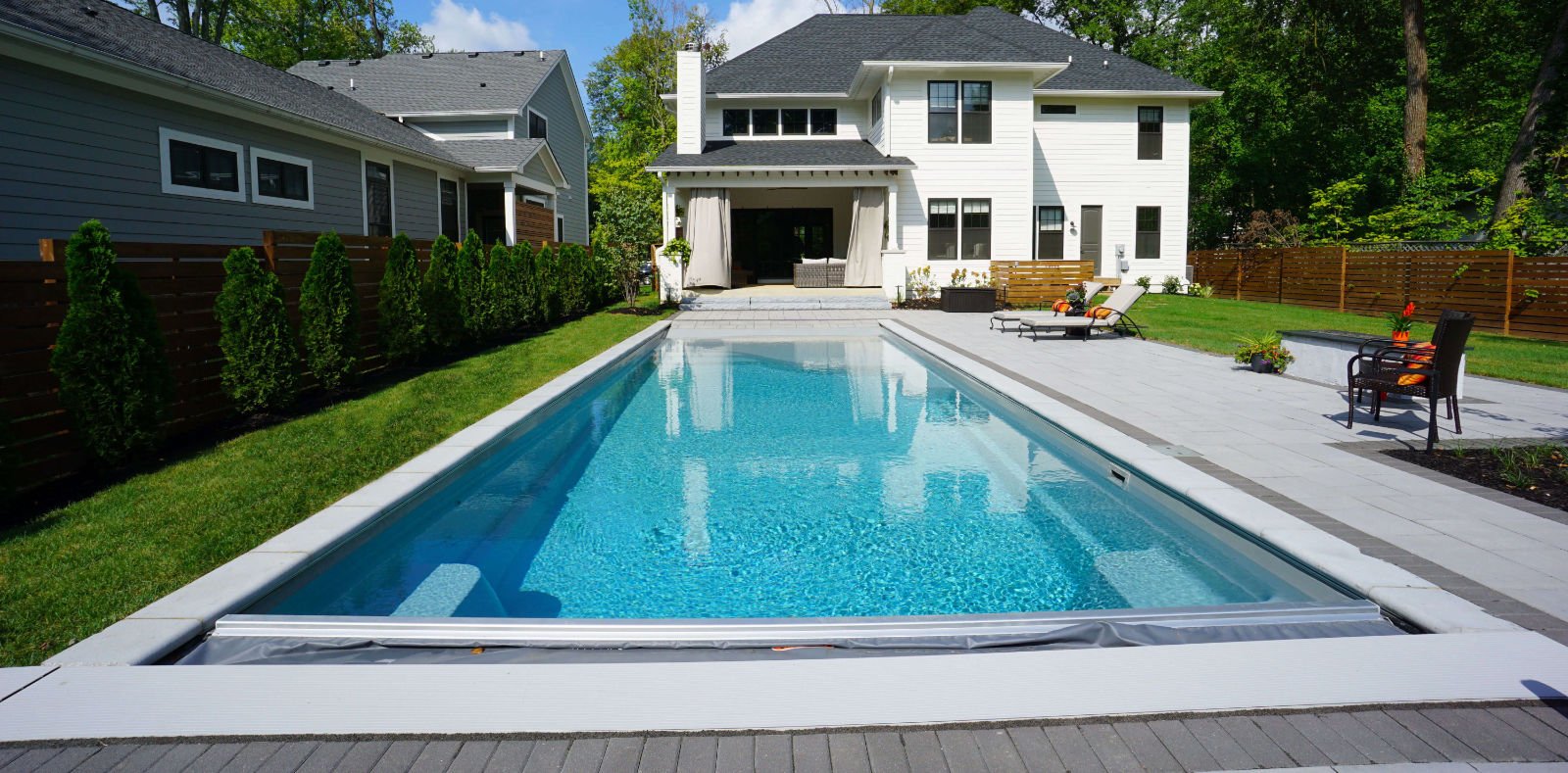 Ground-level view looking down a long rectangular fiberglass swimming pool toward a two-story white modern farmhouse house, featuring an automatic pool cover track system, light gray paver patio, and lawn.