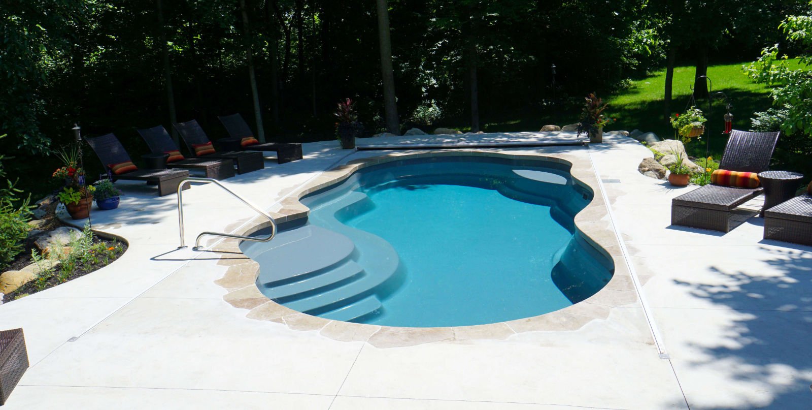 Backyard installation of a Thursday Pools Sun Day freeform fiberglass pool featuring deep teal water, stone flagstone coping, an integrated automatic safety cover, concrete patio, and dark wicker lounge chairs surrounded by trees.