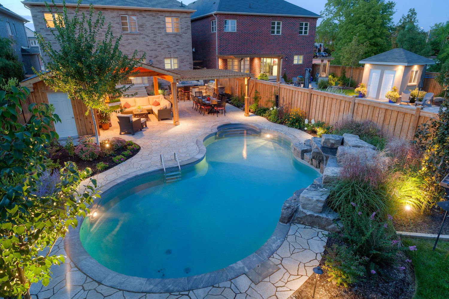 High-angle dusk view of a freeform vinyl liner pool illuminated with soft underwater lighting, featuring dark stone coping, a tiered rock waterfall, flagstone patio decking, and an illuminated outdoor dining and lounge space under a pergola.