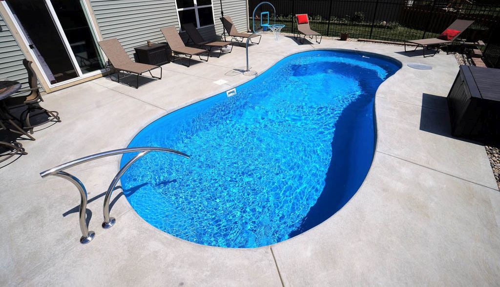Elevated high-angle view of a curved blue fiberglass pool featuring integrated entry steps, a modern stainless steel handrail, surrounding concrete patio decking with lounge chairs, and a poolside basketball hoop.