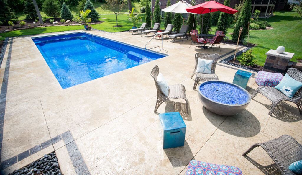 A backyard with a blue swimming pool, lounge chairs, colorful cushions, and a fire pit surrounded by greenery.
