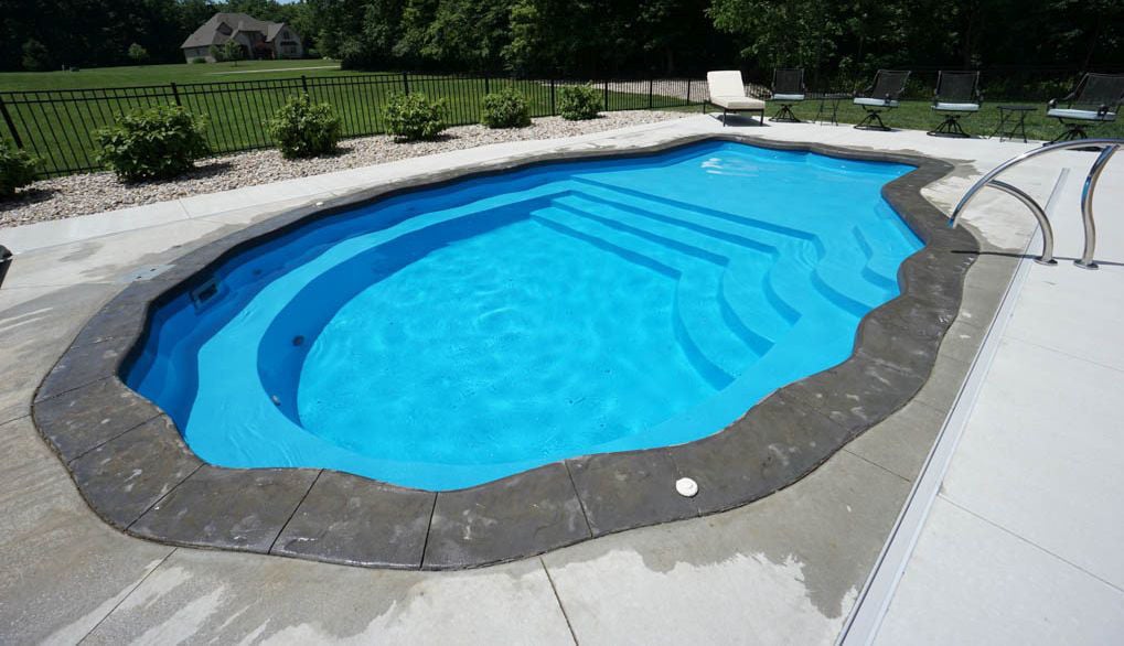 Outdoor installation of a scalloped clam shell-shaped fiberglass pool featuring clear blue water, dark grey stamped concrete cantilever coping, built-in access steps, and a surrounding concrete patio deck.