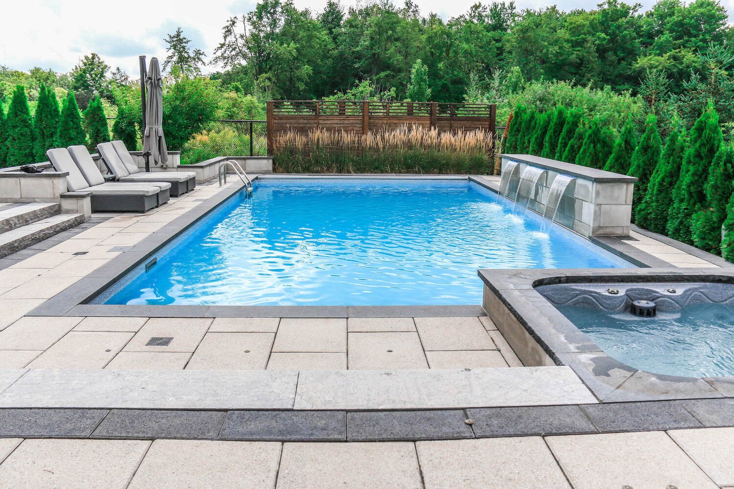 Backyard patio featuring a rectangular vinyl liner pool with bright blue water, a raised stone shear curtain waterfall wall, attached hot tub, and patio lounge chairs.