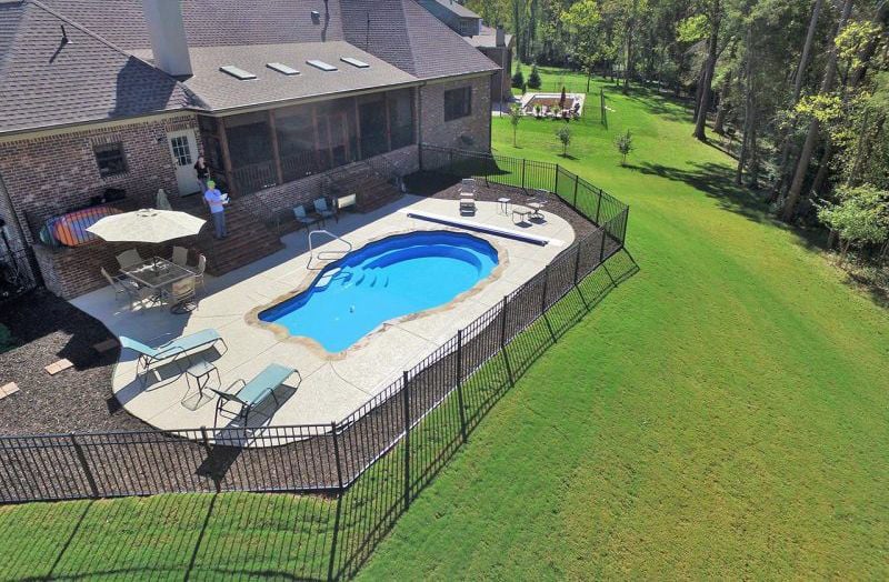 High-angle aerial photograph of a scalloped seashell-shaped fiberglass pool with concrete decking, surrounding black aluminum fence, patio furniture, and a brick residence on a sprawling grassy yard.