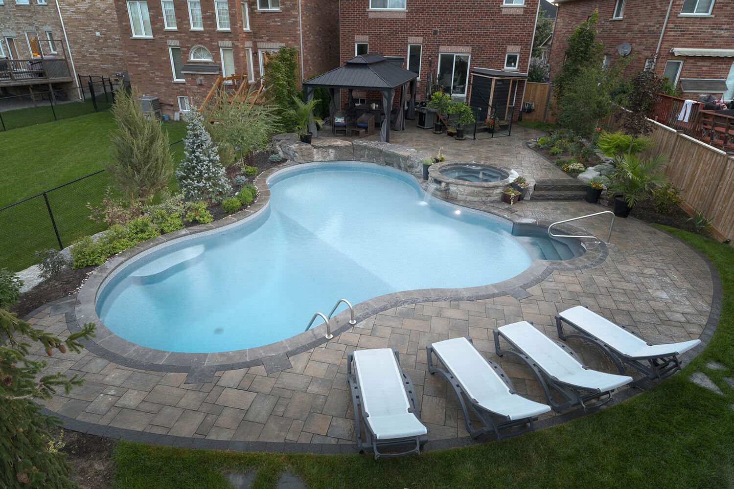 Overhead view of a light blue freeform vinyl liner pool with integrated steps, an elevated circular stone hot tub with a spillover waterfall, paver patio decking, and poolside loungers.