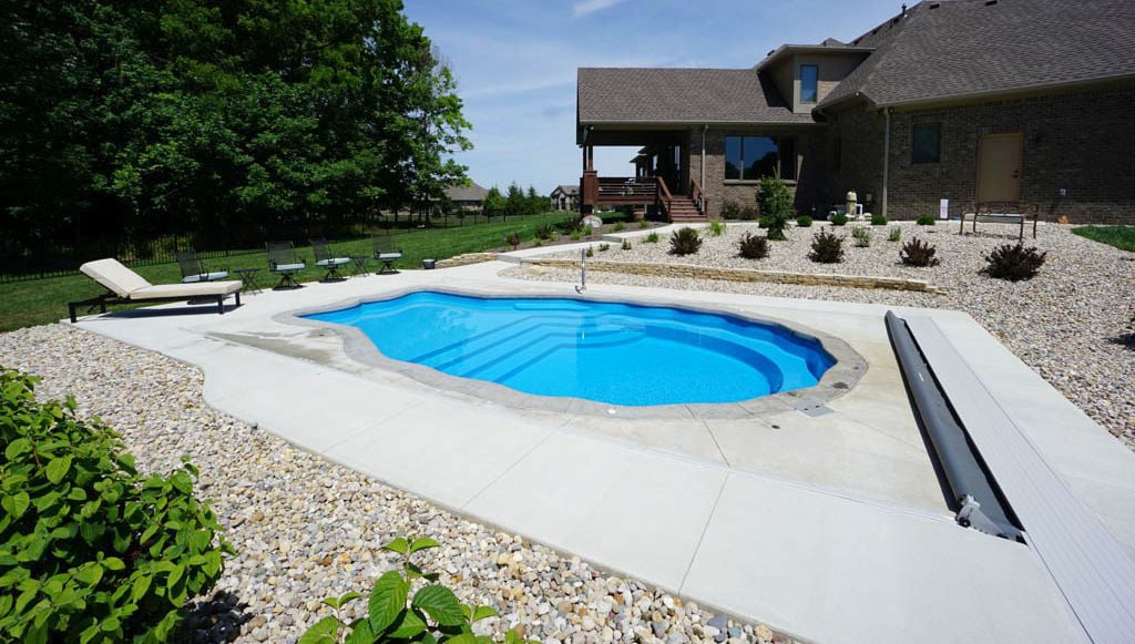 Wide-angle view of a scalloped seashell-shaped fiberglass pool featuring an automatic pool safety cover, concrete patio, gravel landscaping, and lounge seating behind a residential home.