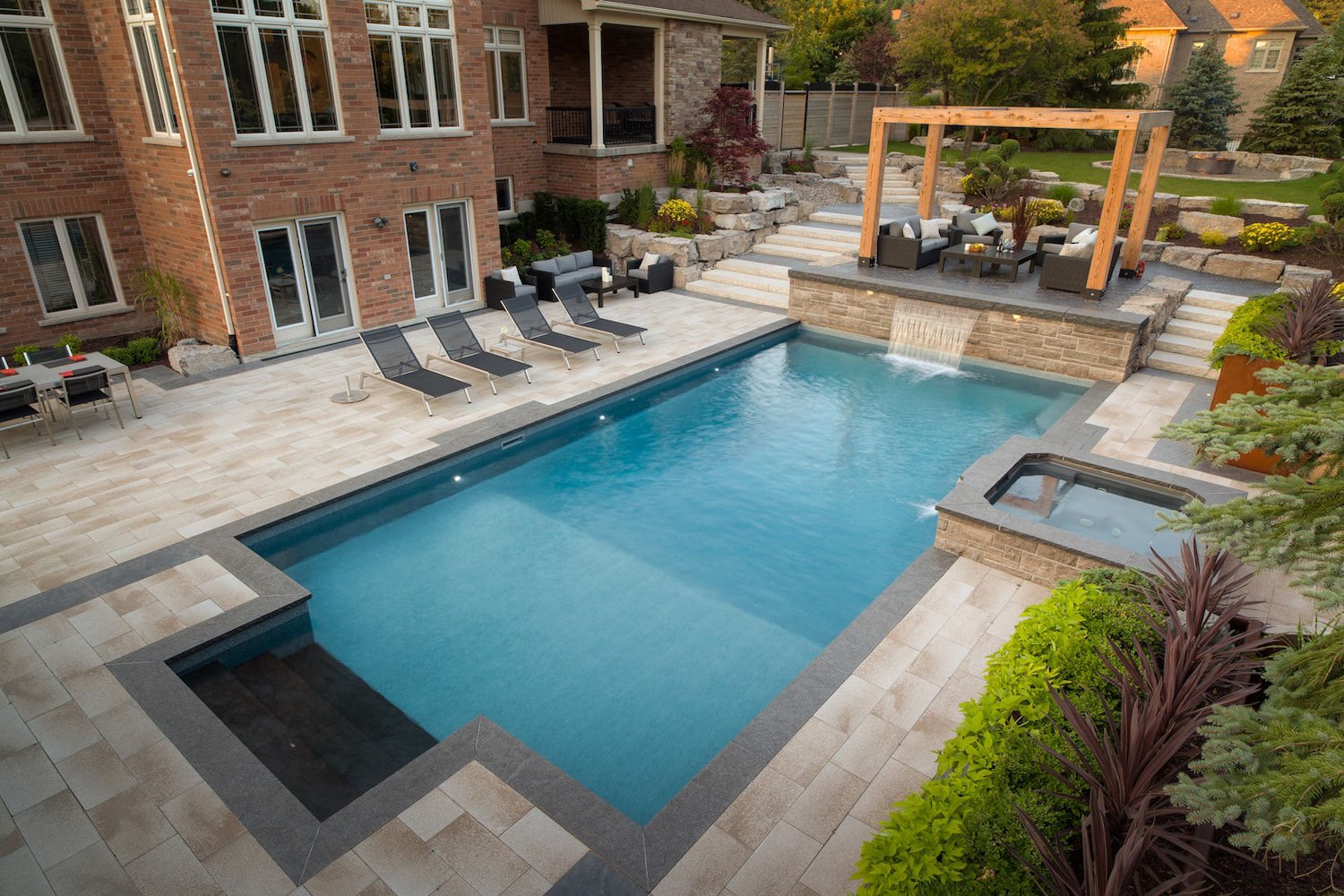 Elevated view of a rectangular vinyl liner pool featuring a dark-tiled tanning ledge/entry steps, a raised stone retaining wall with a shear waterfall, an attached stone hot tub, and surrounding light-colored paver patio with loungers.