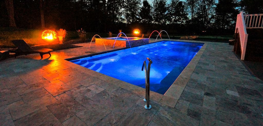 Illuminated swimming pool at night with blue water and stone patio, surrounded by trees and fire features.