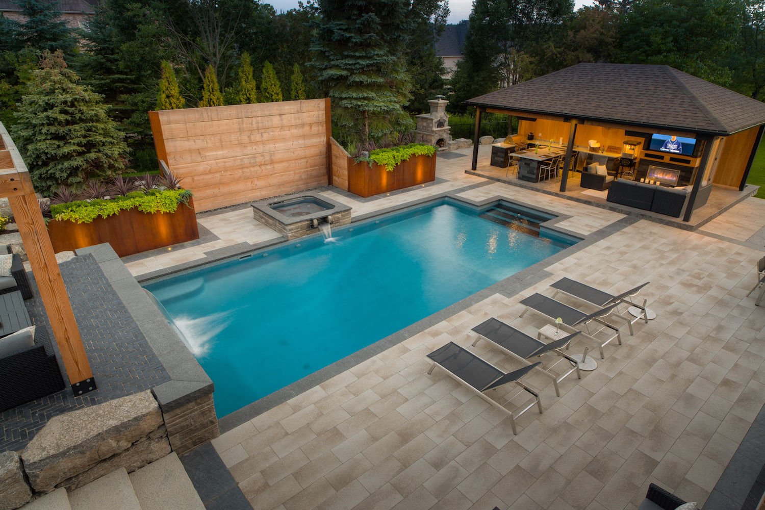 High-angle shot of a sleek rectangular vinyl liner pool featuring an attached square hot tub with a spillover waterfall, a shear curtain waterfall wall, dark stone coping, paver patio, poolside loungers, and an outdoor kitchen area.