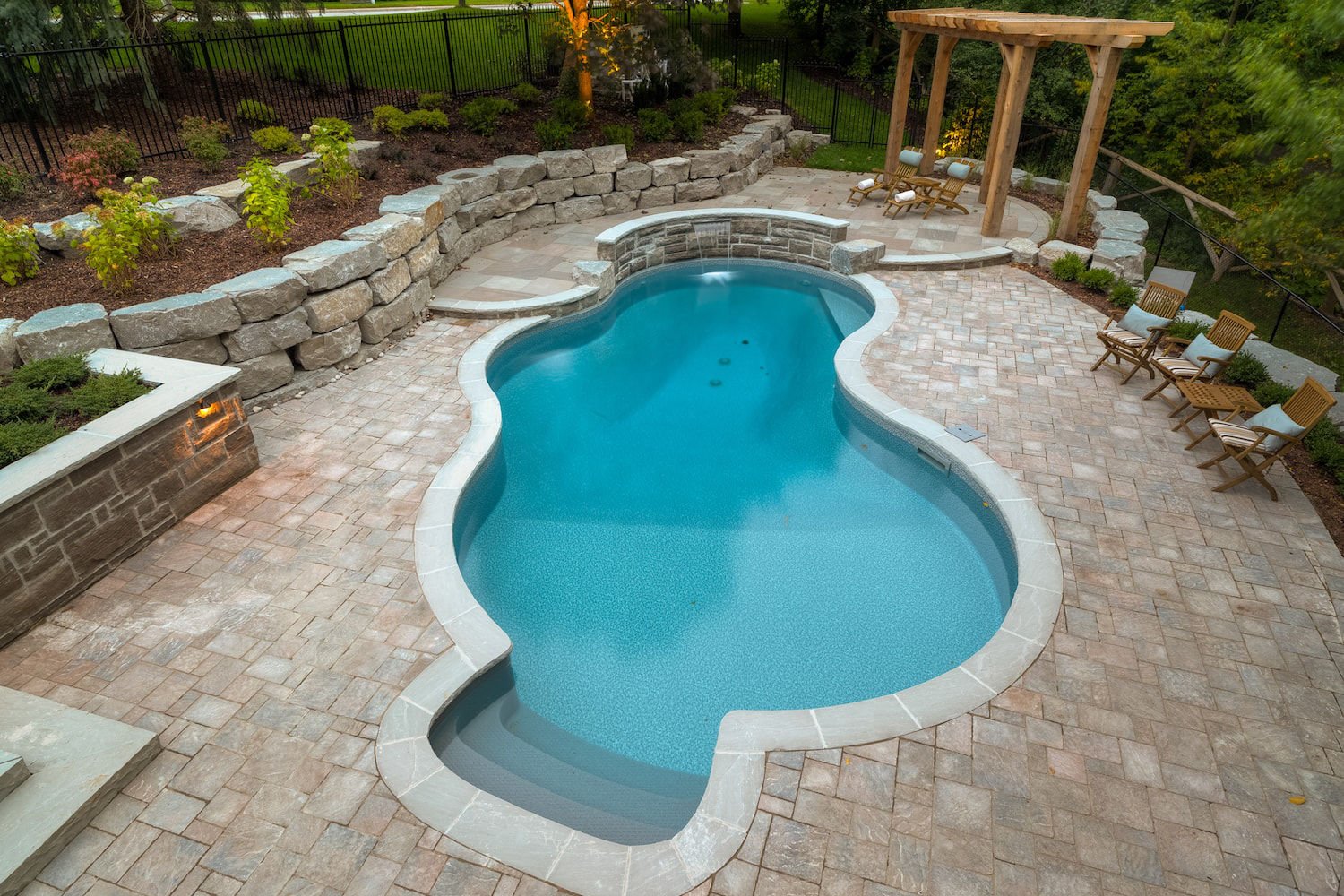 High-angle view of a freeform vinyl liner pool with light stone coping, dark entry steps in an alcove, a curved stone wall with a shear waterfall, stone paver patio decking, and surrounding rock retaining walls.
