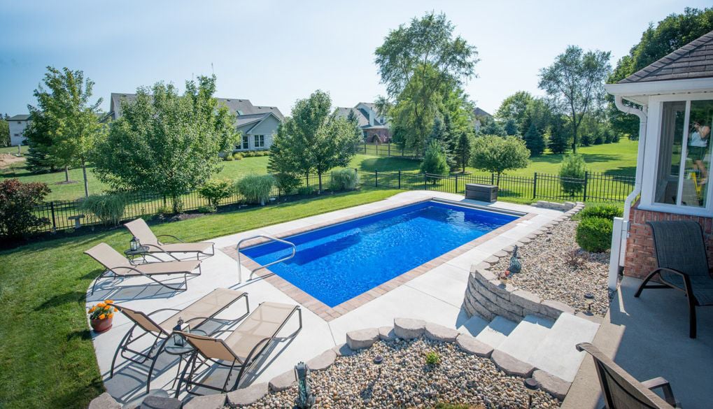 High-angle view of a rectangular blue fiberglass swimming pool with full-width entry steps and stainless steel handrail, surrounded by a light concrete deck with lounge chairs, stone retaining walls, and a black wrought-iron fence around a spacious green y