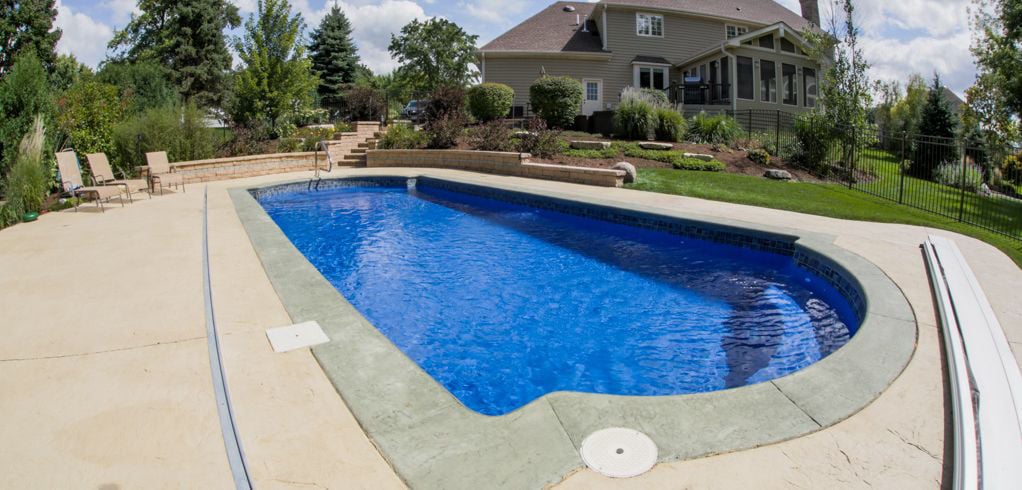 Eye-level view of a Cathedral shaped blue fiberglass pool featuring wide arch entry steps, decorative waterline tile, stamped concrete coping, an automatic safety cover track, and a multi-level retaining wall with landscaping.