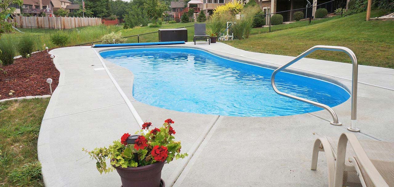 Eye-level view of a freeform curved blue fiberglass pool featuring shallow entry steps, a stainless steel handrail, an automatic cover, concrete deck surrounding, and red mulch landscaping.