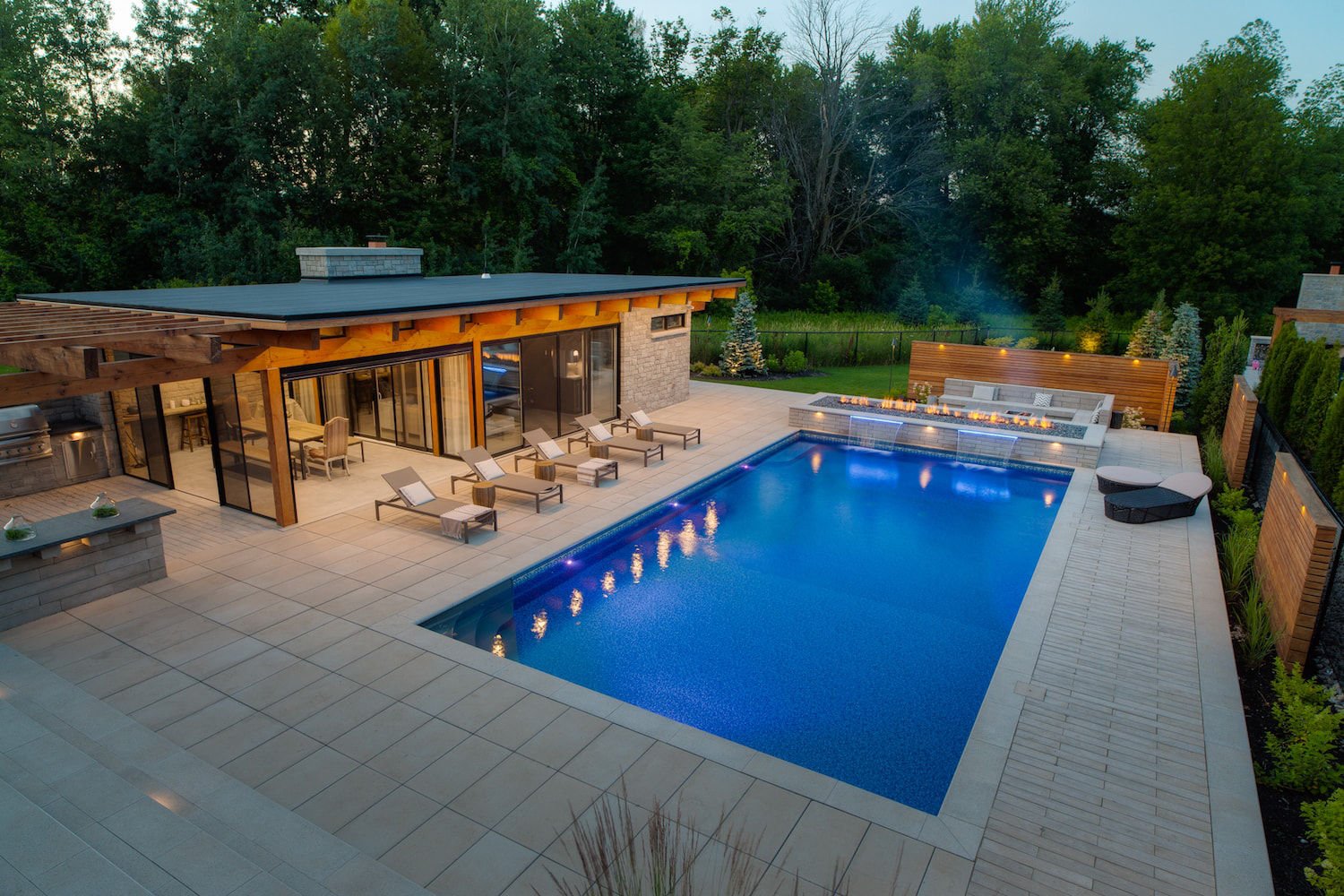 Dusk view of a large rectangular vinyl liner pool illuminated with deep blue LED lights, featuring a linear fire pit with shear curtain waterfalls, poolside lounge chairs, and modern stone patio decking.