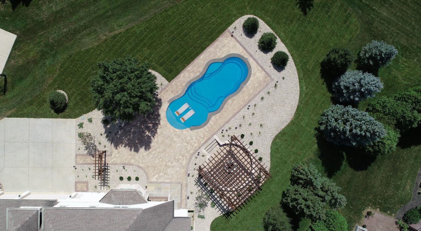 Aerial top-down photograph of a Thursday Pools Sandal Beach Entry freeform fiberglass pool featuring loungers in the beach entry area, a paver patio deck, wooden pergola, and landscaped lawn.