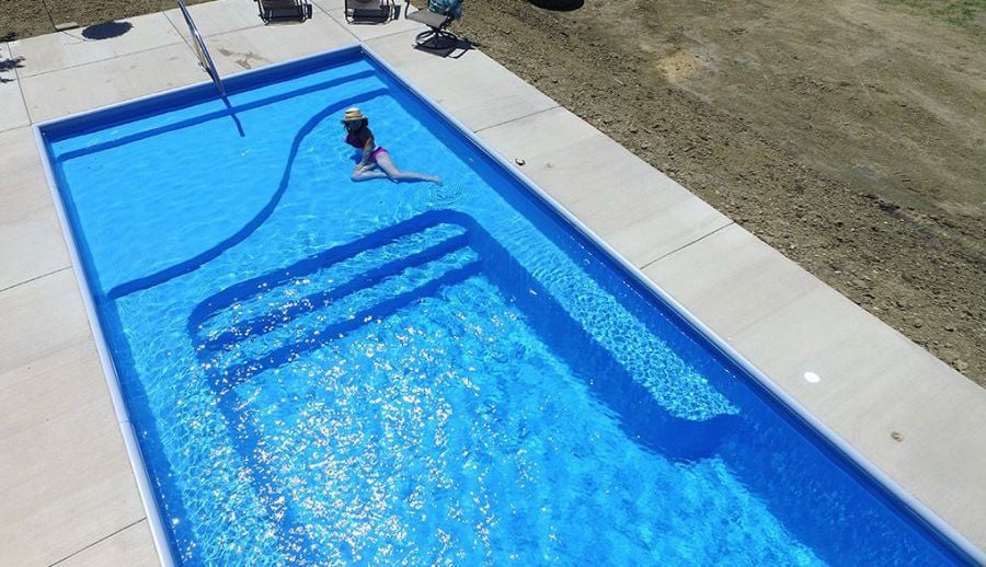 High-angle overhead view of a rectangular blue fiberglass swimming pool featuring an integrated tanning shelf with a woman lounging in shallow water, tiered entry steps, bench seating, and surrounding concrete deck.