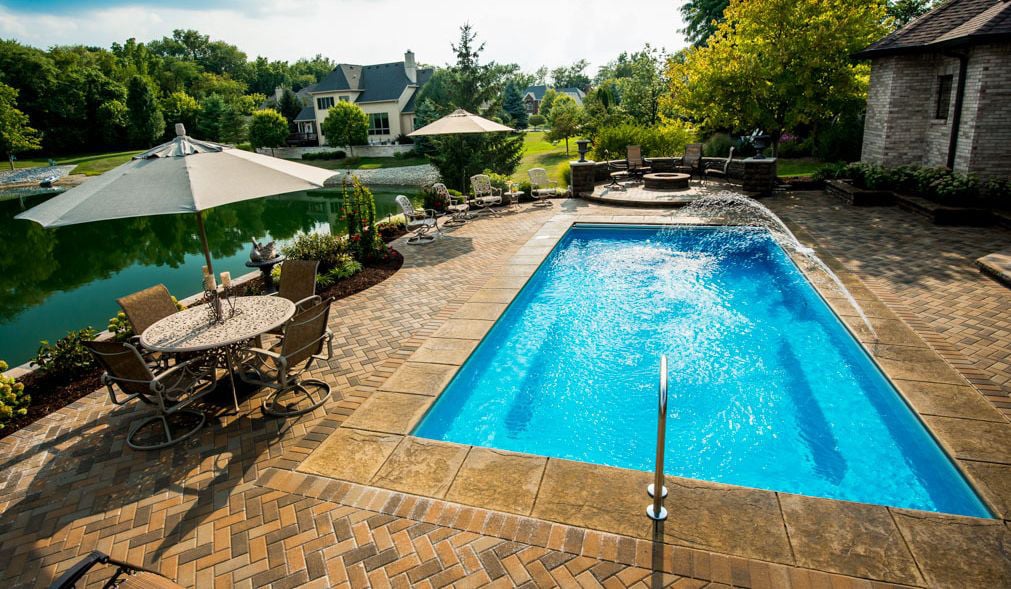 High-angle view of a rectangular blue fiberglass swimming pool with a laminar deck water jet streaming into the water, surrounded by a herringbone stone paver patio, outdoor dining setup, fire pit area, and a scenic pond background.