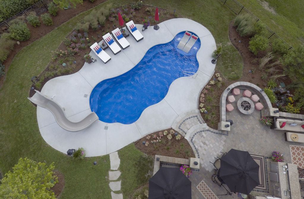 Aerial overhead view of a freeform blue fiberglass pool with a built-in tanning shelf holding two ledge loungers, a curved water slide, concrete decking, and an adjacent stone paver fire pit seating area.