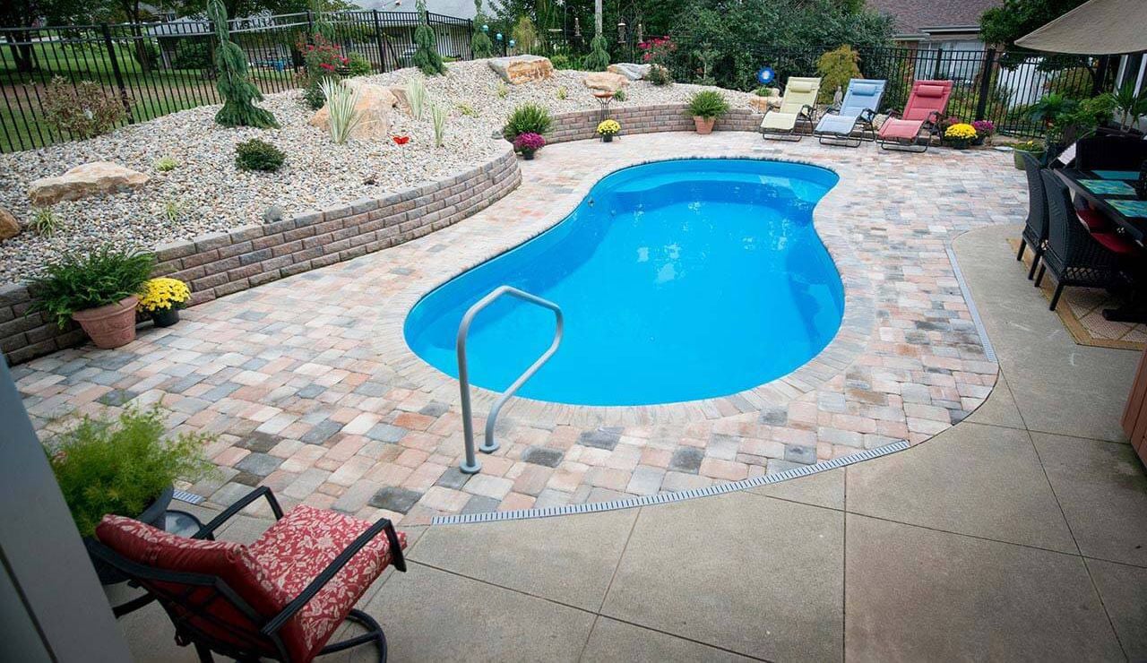 High-angle view of a curved blue fiberglass pool surrounded by multi-color paver decking, grey handrail, stacked stone retaining wall with rock landscaping, and lounge seating area.