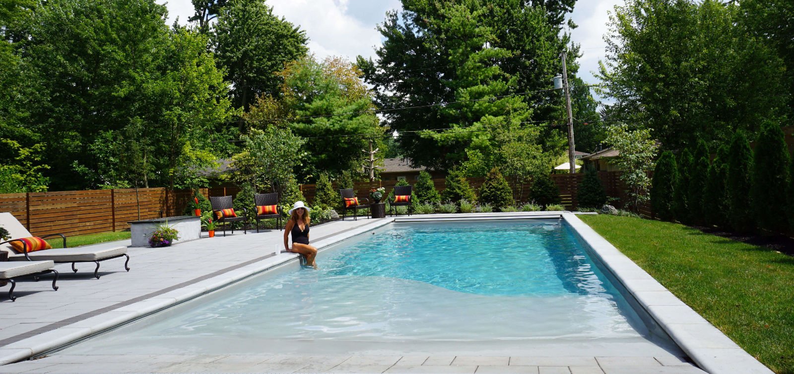 Wide-angle view of a rectangular fiberglass pool with a zero-entry beach slope design, featuring a woman sitting at the pool edge, an automatic pool cover track system, light gray paver patio, patio chairs, and a wooden fence surrounded by trees.