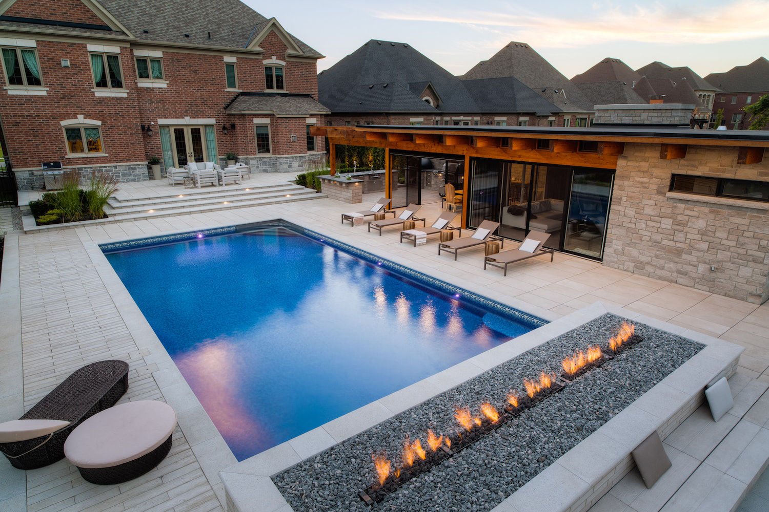 High-angle twilight view of a rectangular vinyl liner pool with vivid blue water, a long linear fire feature set in grey stone gravel, light stone patio pavers, and lounge chairs.