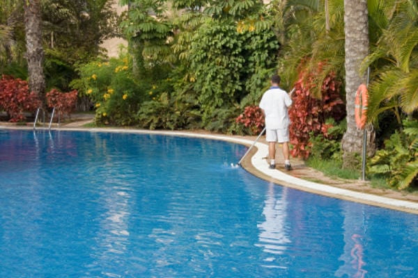 A pool maintenance worker in a white uniform stands poolside using a long pole vacuum to clean the edge of a large outdoor resort swimming pool surrounded by tropical palm trees and lush foliage.