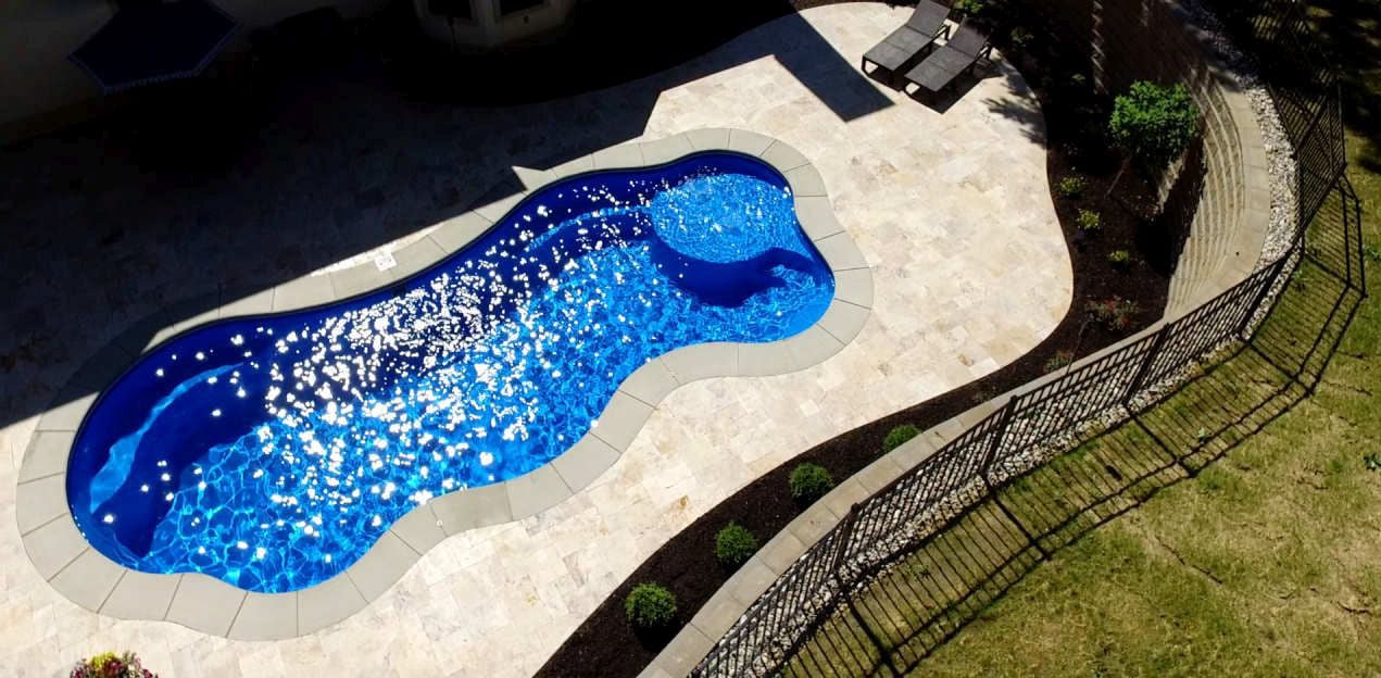 Aerial overhead view of a freeform vibrant blue fiberglass swimming pool featuring an integrated circular tanning ledge, stone coping, travertine paver patio decking, and a curved retaining wall.