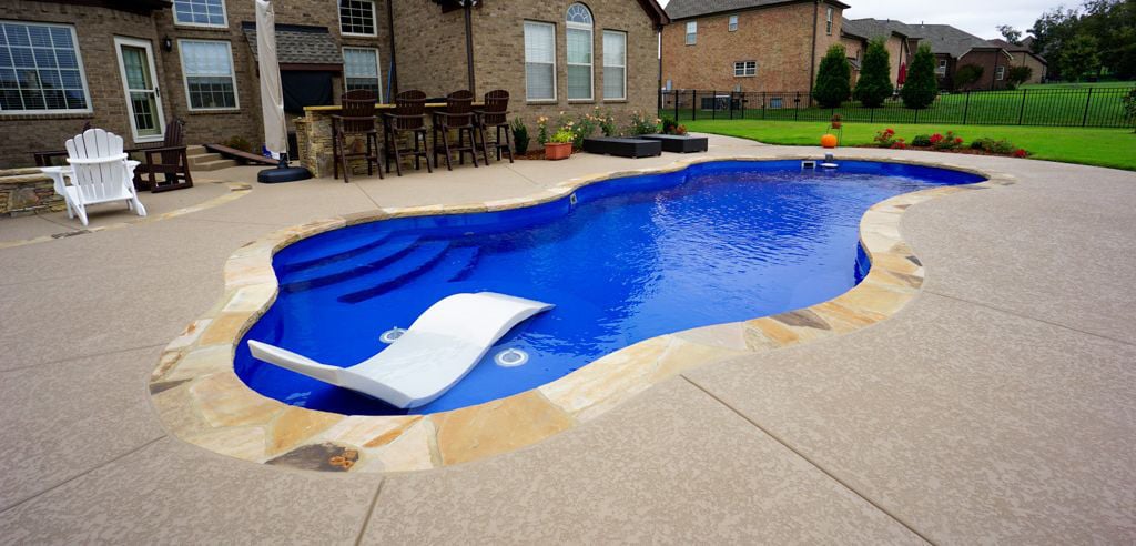 High-angle view of a freeform blue fiberglass pool featuring natural flagstone coping, an integrated circular tanning shelf with a white ledge lounger and bubbler jets, surrounding textured concrete patio, and an outdoor bar area.