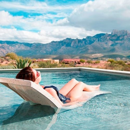 Daytime view of a woman relaxing on a contoured in-pool lounge chair partially submerged on a tanning shelf, overlooking a mountain landscape.