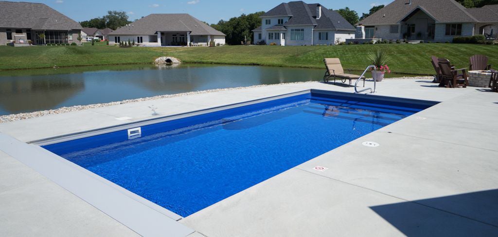 Wide-angle view of a vibrant blue rectangular fiberglass swimming pool featuring full-width entry steps, an automatic pool cover box, and stainless steel handrail, set within a concrete deck overlooking a pond.