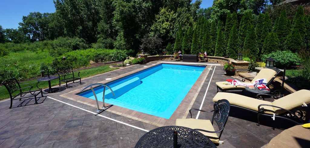 Wide-angle view of a rectangular blue fiberglass pool featuring full-width entry steps, a stainless steel handrail, stamped concrete patio decking, lounge chairs, wrought-iron patio furniture, and a retaining wall with evergreen trees.