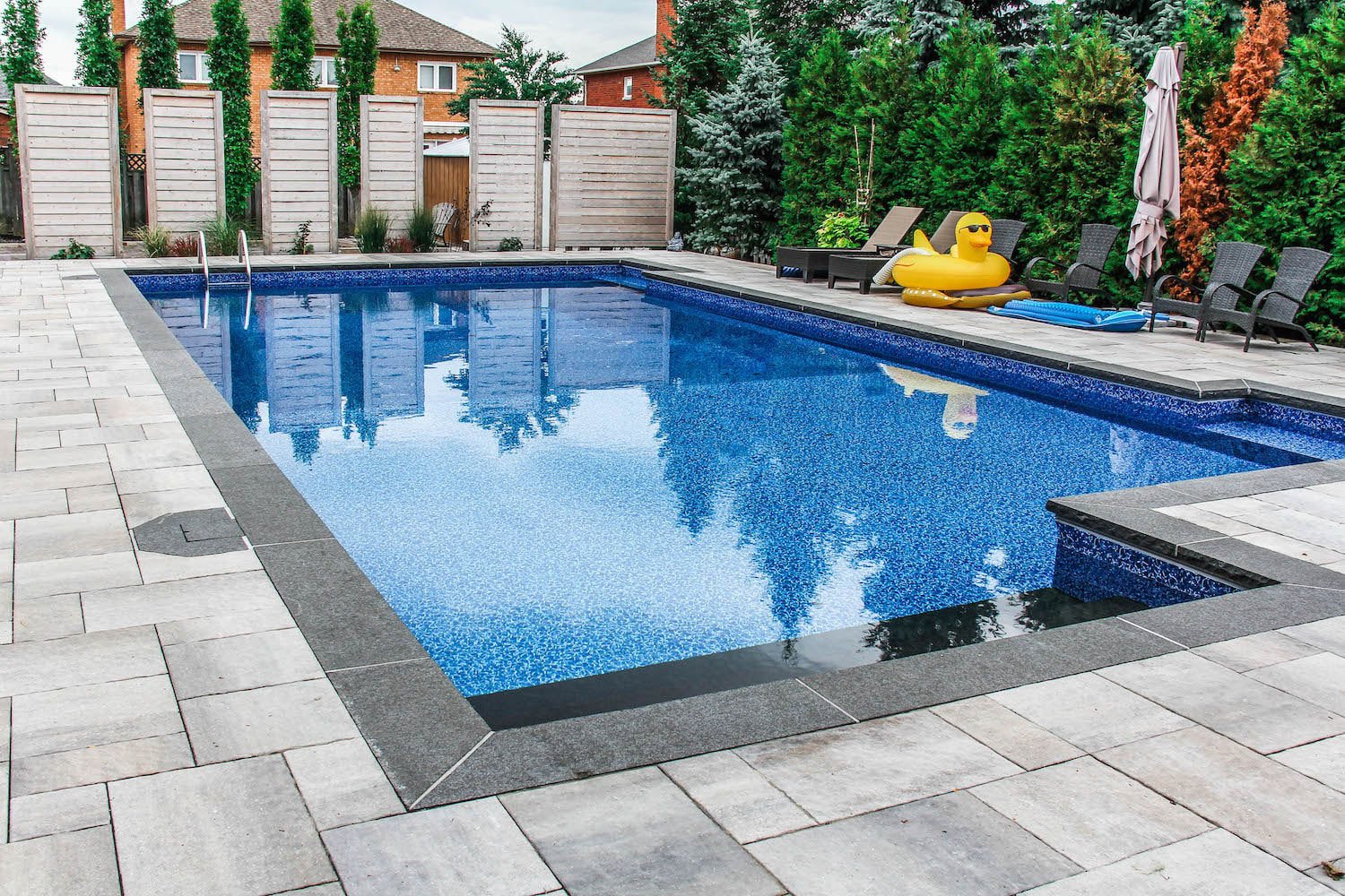 Daytime view of a large rectangular vinyl liner pool featuring vibrant blue mosaic-patterned water, dark coping tiles, light paver decking, entry steps, and poolside lounge chairs with a giant duck float.