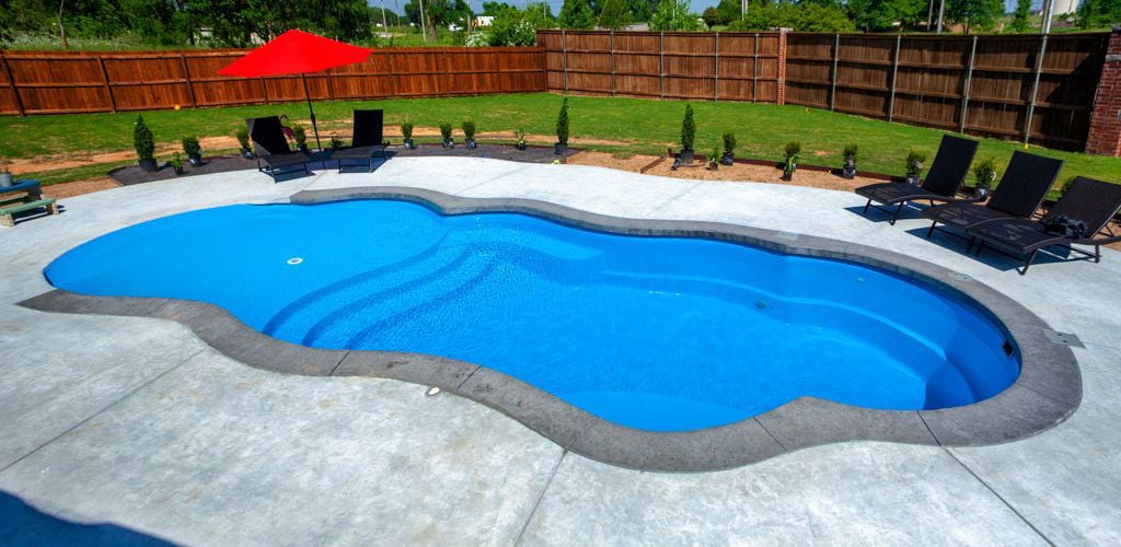 Backyard installation of a Thursday Pools Sandal Beach Entry freeform fiberglass pool featuring bright blue water, zero-entry beach slope, dark grey stamped concrete coping, surrounding concrete patio, and dark lounge chairs with a red umbrella.