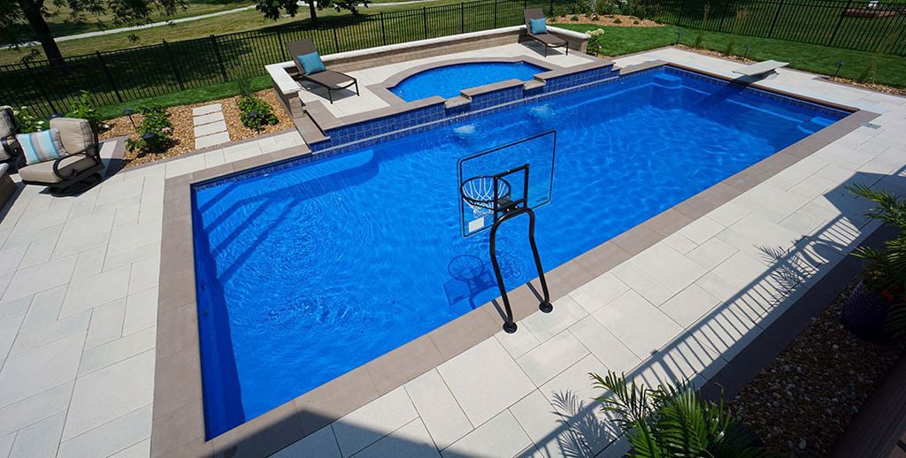 A backyard with a blue swimming pool, a basketball hoop, and lounge chairs beside landscaped greenery.