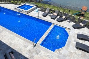 Aerial view of a large luxury backyard stone patio featuring a long blue swimming pool, an elevated square spa with a spillway, an integrated semi-circular splash pad, and dark wicker lounge chairs along the deck.