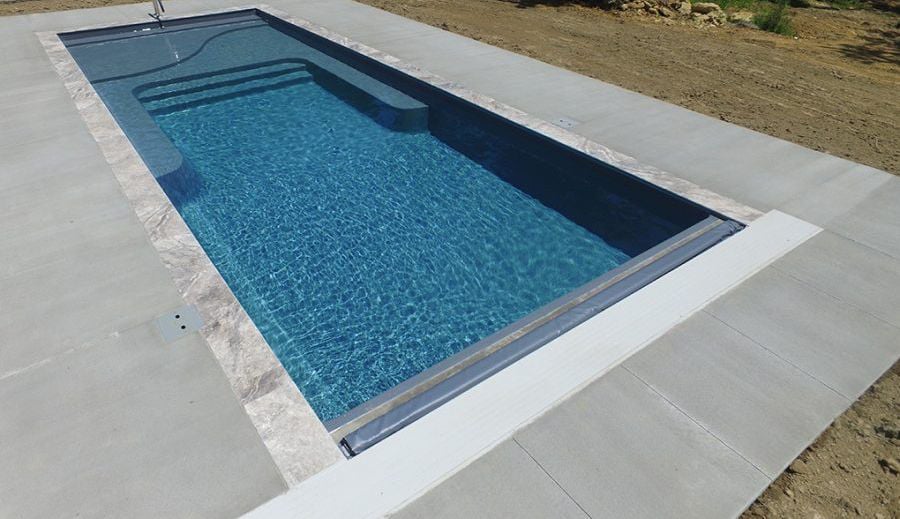 Angled high-angle view of a dark blue rectangular fiberglass pool with an integrated tanning shelf, entry steps, stone coping border, automatic pool cover track housing, and poured concrete decking.