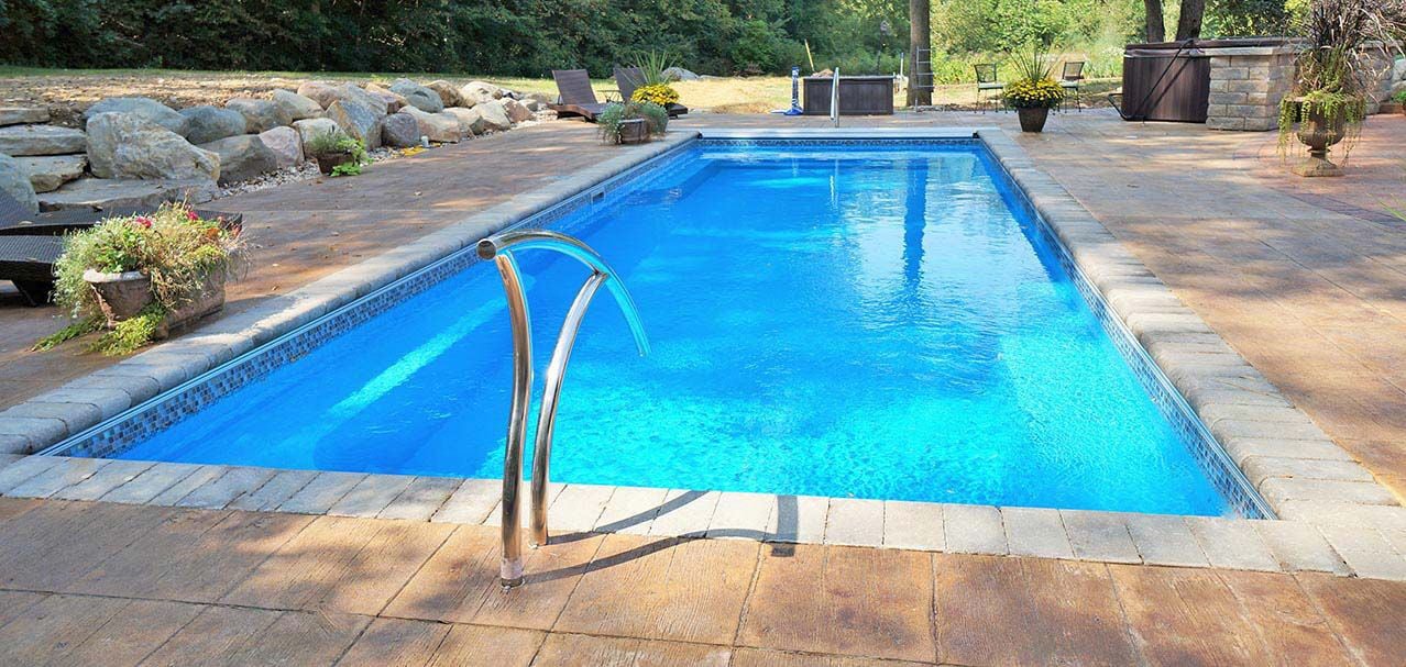 Ground-level length perspective of a rectangular blue fiberglass pool featuring mosaic waterline tile, stone paver coping, a curved stainless steel entry handrail, wood-plank stamped concrete decking, and background boulder landscaping.