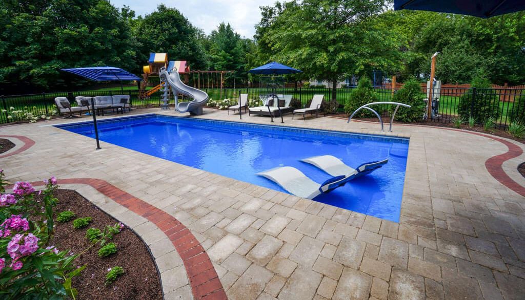 Backyard installation featuring a large rectangular deep-blue fiberglass swimming pool with in-water ledge loungers on an integrated tanning shelf, a curved water slide, lounge chairs under blue patio umbrellas, and paver decking.