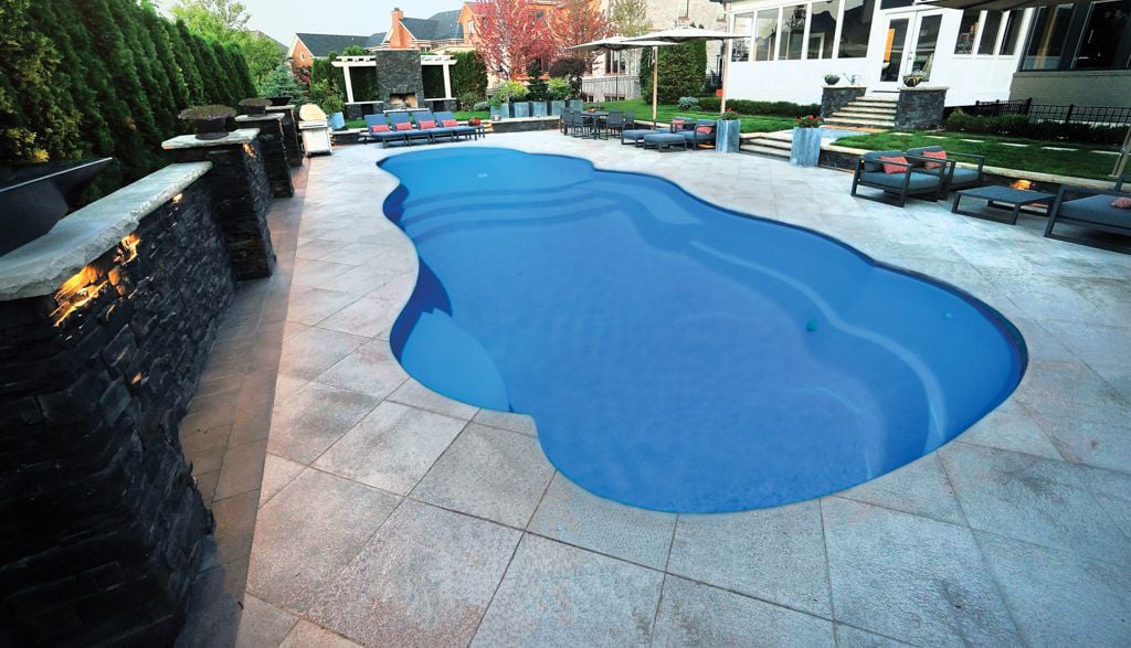Ground-level view of a Thursday Pools Sandal Beach Entry freeform fiberglass pool featuring rich blue water, stone paver patio decking, a stacked stone retaining wall with integrated lighting, and outdoor lounge seating.