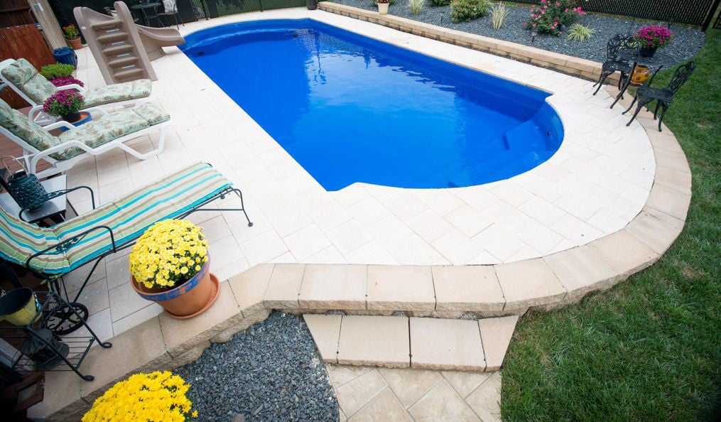 Elevated view of a Cathedral shaped blue fiberglass pool featuring light tan paver patio decking, a poolside water slide, lounge chairs, potted flowers, and a block retaining wall edge.