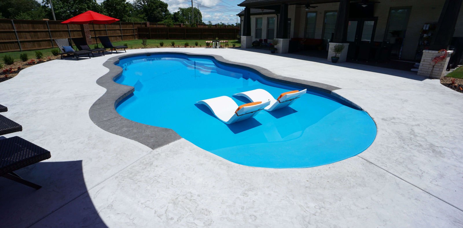 Backyard installation of a Thursday Pools Sandal Beach Entry freeform fiberglass pool featuring bright blue water, zero-entry beach slope, dark grey stamped concrete coping, surrounding concrete patio, and dark lounge chairs with a red umbrella.